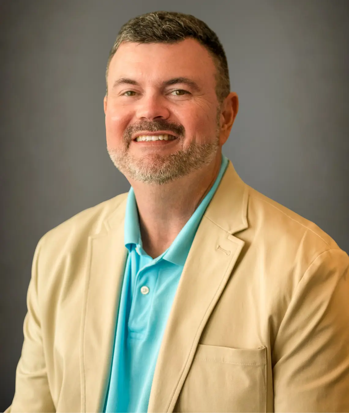 Middle-aged man with beard smiling, wearing tan suit jacket and turquoise collared shirt