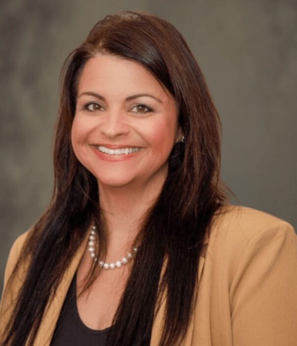 Smiling woman with long brown hair wearing tan blazer and pearl necklace, professional headshot
