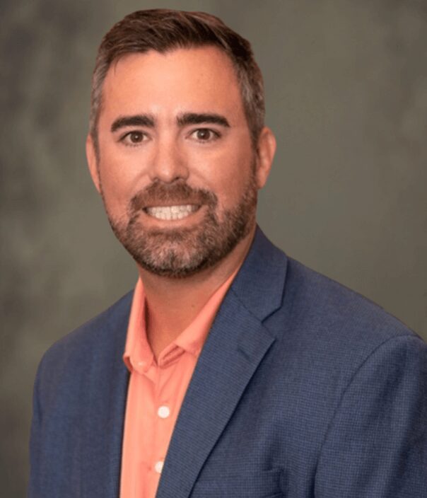 Smiling businessman in blue suit jacket and orange collared shirt against gray background, headshot portrait