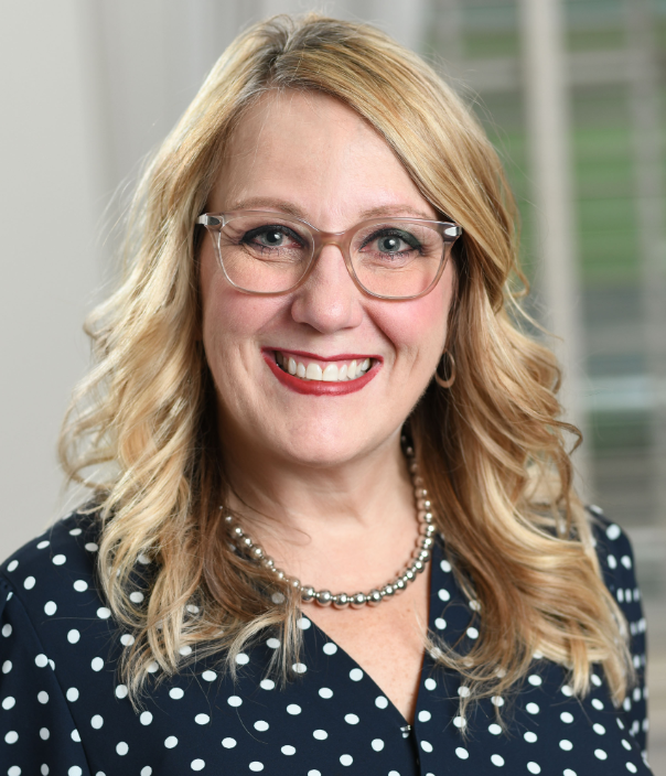 Smiling blonde woman with glasses wearing polka dot blouse and pearl necklace, business professional headshot